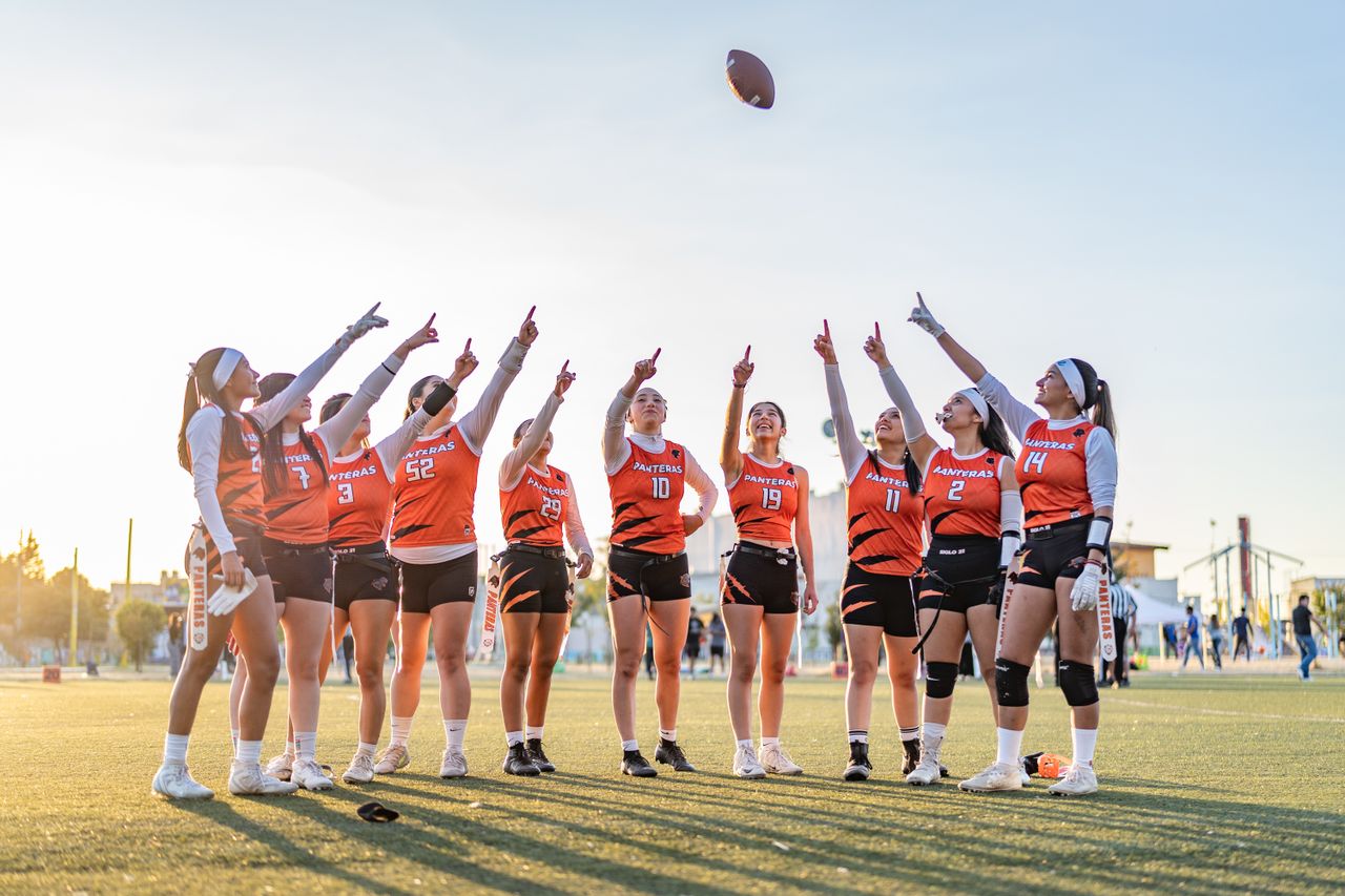 A women's flag football team raises their fingers in celebration on a sunny day.
